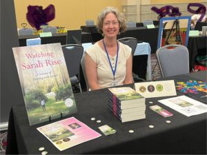 Jenny is sitting at a table with a poster of her book cover and a stack of her books in front of her. She is wearing a cream colored tank top and a lanyard with a name tag. She is smiling. 