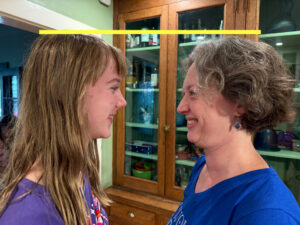 Amy and Jenny are facing each other and smiling. The picture is a close up of their heads in side profile. There is a yellow line added to the photo crossing from Amy’s head to Jenny’s and showing that Amy is taller than Jenny