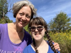 Jenny in a purple t shirt and Sarah in a blue t shirt, smiling. Jenny has her arm around Sarah’s shoulders