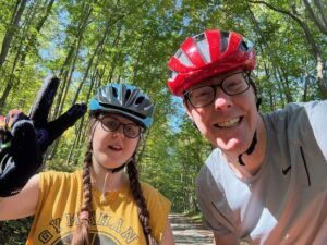 Sarah in a blue bike helmet and yellow tank top; Carl in a red bike helmet and gray shirt. They are both smiling and Sarah is making a peace sign. 