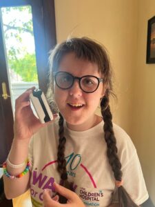 Sarah standing in a hallway with a tall window behind her and a beige wall to one side. Her hair is in braids and she is wearing a white t-shirt and glasses. She is holding a black and white cassette player up near her cheek and smiling