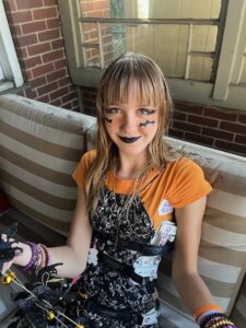 Amy sitting on a beige  and white striped porch swing. She is wearing an orange shirt under a black and white dress and her black and white Whisper brace for scoliosis. She has orange eye shadow and cheek shadow and black bats drawn on her cheeks and near her eyes.