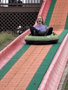 Amy in a lavender sweater and black leggings, leaning back in an inner tube as she goes down a wide pink, green, and orange striped slide. Her mouth is open in a smile.