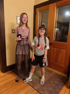 Amy and Sarah standing in their entryway room next to the front doors of their house. Amy is wearing a pink shirt and shorts on top of fish-net tights and with a black mesh dress over everything. Sarah is in black and white Addidas shorts with a gray St. Therese Tigers shirt.