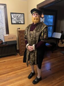 Carl is wearing a black hat, fake beard, and fancy clothes a la Henry VIII