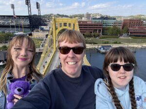Amy, Carl, and Sarah look like they are very high above a yellow bridge. They are at the top of a Ferris Wheel with PNC Park behind them. They are smiling. Carl and Sarah have sunglasses on. 