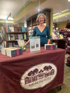 Jenny in a teal long-sleeved v-neck shirt standing behind a table draped in a maroon tablecloth with the Swarthmore College logo on the front of the tablecloth. The table has stacks of Watching Sarah Rise and a sign about the author talk