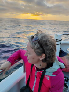 Jenny in a pink raincoat looking over the side of a boat at the ocean with a bit of sunset in the background. The picture is taken from above and Jenny's face is in profile.