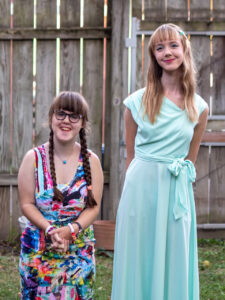 Sarah and Amy standing outside with a tall gray fence behind them. Sarah is in a multi-colored sleeveless dress and Amy is in a sea-foam colored dress with a bow at the waist. Sarah’s hair is in two braids. Amy’s hair is down and loose. 