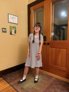 Sarah in a black and white dress, white socks and black shoes, standing inside in front of two doors.