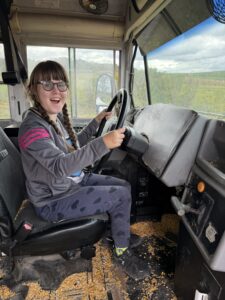 Sarah wearing blue sweatpants and a gray sweatshirt, sitting in the driver's seat of a stationary firetruck and smiling hugely.