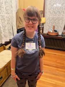 Sarah standing in a front entry-room to her house, wearing a blue s-shirt with musical notes on it and a sign taped to her that reads "I hate music."