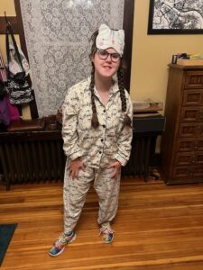 Sarah standing on a wooden floor with a wooden dresser behind her and a white lace curtain on the window behind her. She is wearing white flannel long-sleeved and long-pant pajamas with music notes all over them. Here hair is in braids and she has a white cat sleep mask on her forehead.