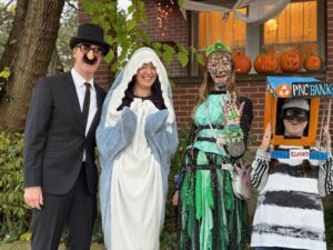 Carl, Jenny, Amy, and Sarah are standing in front of their porch with a tree behind them. Carl is wearing a top-hat, suit, and Groucho Marx nose and glasses with bushy eyebrows. Jenny is wearing a fuzzy shark blanket with flippers. Amy is wearing layers of green with a green tiara and her Whisper Brace. Her makeup looks green and metallic and covers her face and left hand. Sarah is wearing a shirt with black and white horizontal stripes, blue pants, and a cardboard box painted as PNC bank on her head.
