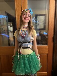 Amy wearing a yellow shirt under a small blue sweater vest and a green tutu. She has many colored necklaces and bracelets and hair clips and an array of brightly colored gems across her cheeks