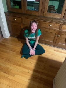 Amy is sitting criss-cross applesauce on a hard-wood floor with wooden cabinets behind her. She is wearing a green shirt and green pants, with a huge laughing smile on her face and her eyes closed.