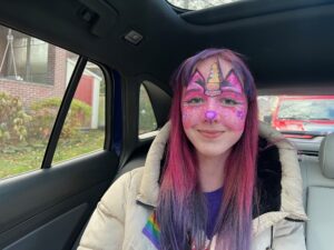Amy sitting in a car with a white winter coat and pink hair with purple tips and a pink purple unicorn cat depicted in face paint