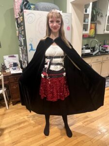 Amy is wearing a white shirt and red sequined knee-length skirt over black tights. she has black stripes across her torso from her Whisper Brace straps. She is wearing a long black cloak and has red eye shadow, a black bat drawn on her cheek, and a trickle of lipstick blood coming out of her red lips.