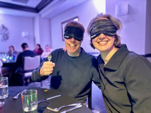 Carl and Jenny are sitting at a table in a dimly purple-lit room, both in black tops, and with black eye masks on (the kind people wear for sleeping). They are smiling and Carl is holding a fork.