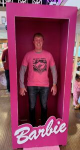 Carl holding himself rigidly to mimic a Ken doll, standing in a giant Barbie Box, while wearing a pink t-shirt and blue jeans.