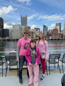 Carl, Sarah, and Amy are all wearing pink and standing on the deck of a boat with downtown Pittsburgh in the background behind the river