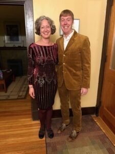 Jenny in a maroon long-sleeved, knee-length dress with vertical stripes of metallic gold; Carl is in a brown corduroy suit (matching jacket and pants) over a white shirt. They are standing next to their front door.
