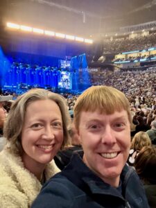 Jenny with a fuzzy white sweater and Carl with a black fleece. Mainly you just see their smiling faces with a blue-lit stage far in the background and a crowd of people in the stands of the concert arena