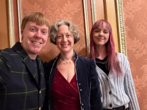 Carl, Jenny, and Amy are standing in front of a brown-ish wall at the Benedum, a venue for musicals. Carl is in a plaid coat, Jenny is in a red velvet dress and blue overcoat. Amy has a dark blue top and light blue sweater.