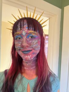 Amy with pink hair and face/neck paint that makes her look like a mermaid (pink, purple, white, and blue). She is wearing a tiara from the SIX musical.