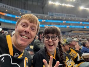Carl and Sarah in Penguins shirts, smiling broadly with an arena full of Penguins fans behind them.