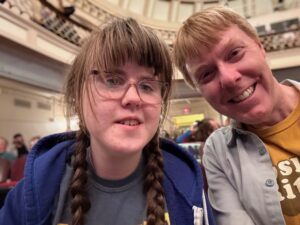 Sarah wearing a grey t-shirt, blue sweatshirt that is unzipped, and glasses, smiling next to Carl, who is wearing a grey button shirt open over a yellow Josh Ritter shirt. There is a balcony behind them.