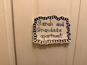 A hand-written sign saying "Sarah and Grandad's apartment" with a black and white boarder around the words, is taped to a white door