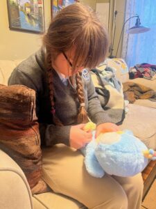 Sarah in khaki pants and a grey v-neck school uniform sweater is sitting on a beige sofa holding a light blue penguin stuffed animal and looking down at it.