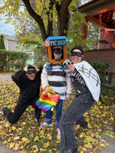 Anna's partner and Anna are dressed in black with black eye masks. Anna is holding a large white pillowcase with a black $ painted on it. Sarah is in a black and white shirt with a black eye mask. She is holding a rainbow striped back for trick or treating. She has a cardboard PNC bank on her head.