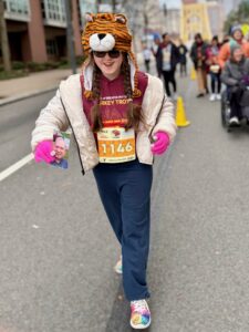 Sarah smiling and looking at the ground ahead of her as she does the Turkey Trot. She is wearing a white coat, pink gloves, a brown turkey trot shirt, and a hat that looks like a tiger head.
