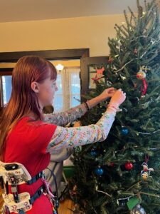 Side view of Amy wearing a red Christmas shirt under her Whisper brace while she puts an ornament on the Christmas tree