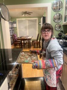 Sarah in a blue sweatshirt and red pants and white apron holding a tray of gingerbread cookies with her hands in oven mitts