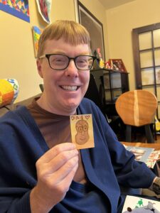 Carl smiling while holding up a small watercolor art piece made by Amy of a potato with a face 