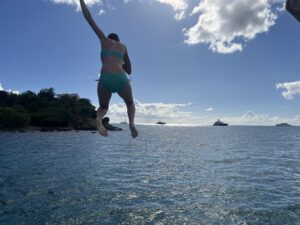 A view of the ocean and blue sky with Amy seen from behind as she sails through the air after jumping off the top of a sailboat. 