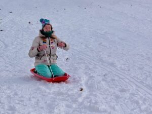Amy kneeling on a pink sled, dressed in teal snow pants and a white winter coat she decorated with fabric paint, pink gloves, and a blue hat. She is on a snowy hill 