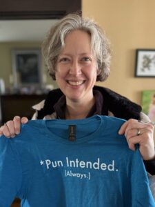 Jenny smiling hugely while holding a blue t-shirt in front of her. She got the shirt for Xmas and it says “*Pun Intended. *Always”