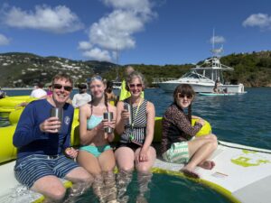 Carl, Amy, Jenny, and Sarah in swimsuits and holding metal drink tumblers while sitting on a crescent shaped raft on a sunny day with a blue sky and wispy white clouds