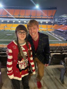 Sarah and Carl standing and smiling with an empty football stadium behind them. Sarah is in a red and white Christmas sweater with a penguin on the front. Carl is in a red shirt, khaki pants, and green/blue/yellow plaid jacket