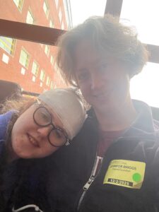 Sarah with her head wrapped in white gauze to keep the EEG leads tidy, leaning against Jenny as they sit in their hospital room for a routine EEG
