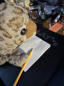 Close-up of a bobcat stuffed animal looking like it is holding a pencil and standing on a piece of paper with a rough paw drawing on the paper