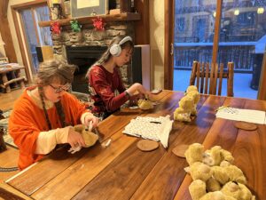 Sarah in an orange shirt and Amy in a red shirt, seen from the side as they sit at a wooden table. They are assembling small plastic scoliosis braces. there is a pile of small beige stuffed animal bears on the table. 