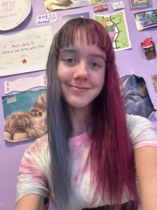 Amy sitting in her bedroom with pictures of cute cats behind her. The right half of her hair is blue and the left half is pink. She is wearing a tie dyed shirt. 