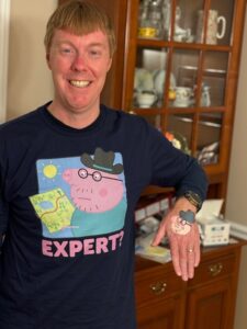 Carl smiling while wearing a dark blue tshirt that has Daddy Pig (from Peppa Pig) holding a map. Under the picture it reads “expert?”  Carl is holding. His left hand next to the shirt so the painting Amy did of Daddy Pig is visible. 