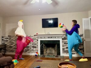 Jenny and Amy are facing each other in a spacious living room. Jenny is wearing a fuzzy white goose robe that has the goose head as a hood over her head. She is wearing huge pink pants from a yard game. Amy is wearing a purple sweater and huge blue pants and yellow goose foot slippers. Amy is tossing a beach ball towards Jenny
