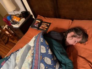 Sarah is sleeping on her side on orangey-brown sheets with a blue blanket. Next to her on the pillow is a small photobook spread open so two photos of her with her Granddad are visible